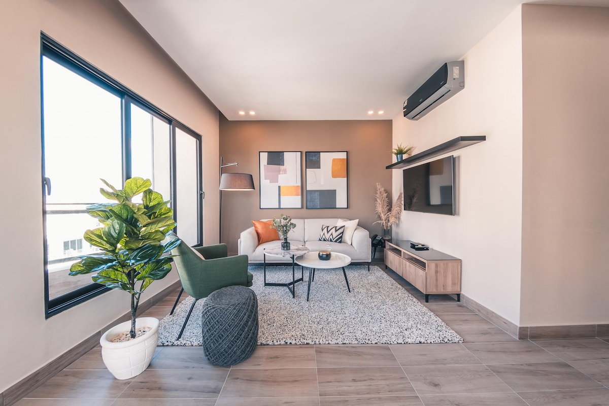 A modern living room showcases large windows allowing natural light to fill the space. Comfortable seating includes a white sofa and green accent chairs. A stylish coffee table is placed on a textured rug, accompanied by a decorative plant and wall art that adds character to the room.