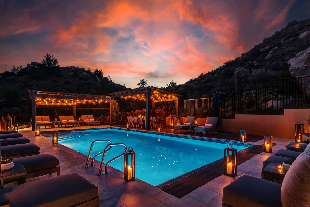 An outdoor pool is illuminated by soft lighting against a vibrant sunset sky. Comfortable lounge chairs surround the sparkling blue water, and lanterns line the poolside, creating an inviting ambiance for relaxation. The area is framed by hills in the background.