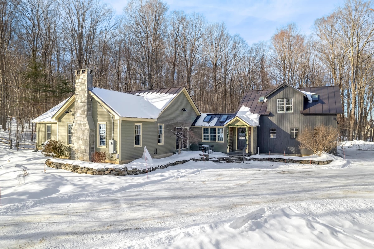This exterior view features a spacious four-bedroom home set in a snow-covered landscape. The building showcases a mix of stone and wooden siding, complemented by a metal roof. A stone wall borders the entrance, creating a welcoming approach surrounded by trees.