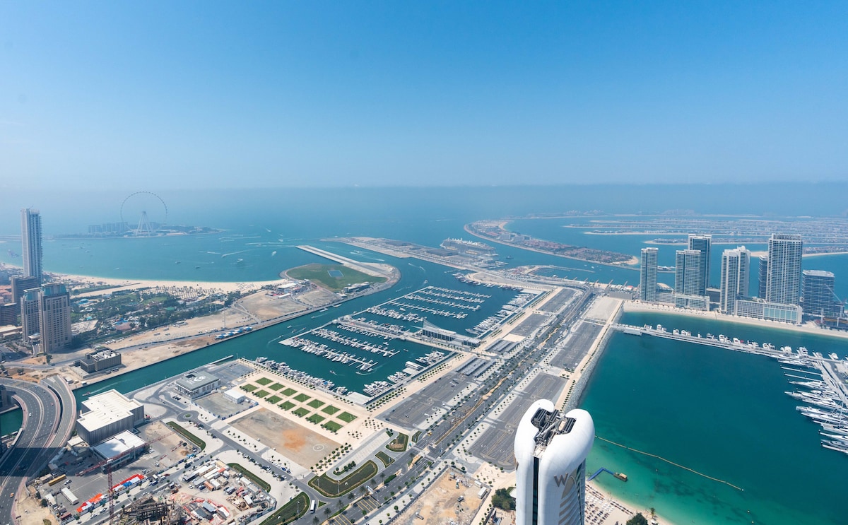 A panoramic view of the coastline features the sea and marina in vibrant shades of blue. The Dubai Eye stands prominently in the distance, surrounded by modern skyscrapers and lush green areas. The shoreline is dotted with yachts and vibrant waterfront spaces.
