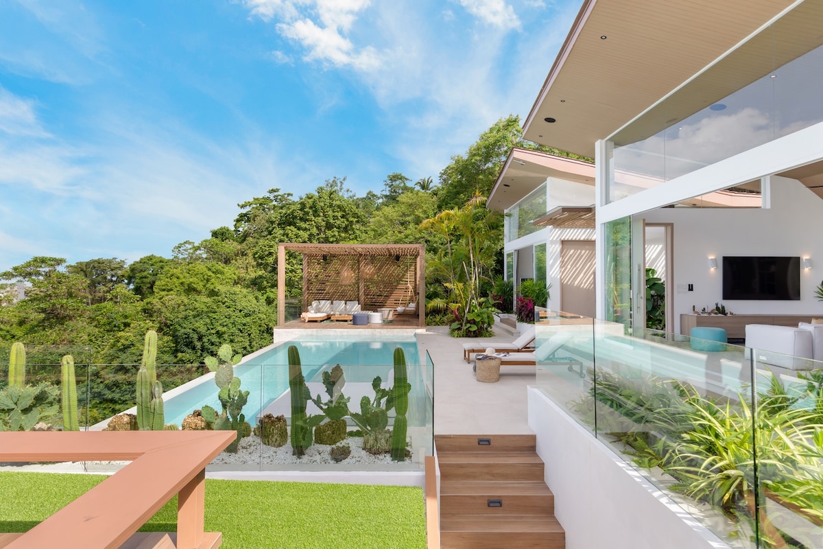 The villa's outdoor living space features an inviting infinity pool surrounded by landscaped gardens, including several tall cacti and greenery. A stylish wooden table is positioned nearby, with a shaded area providing a comfortable lounge space. Expansive glass walls allow for integration with the lush surroundings.