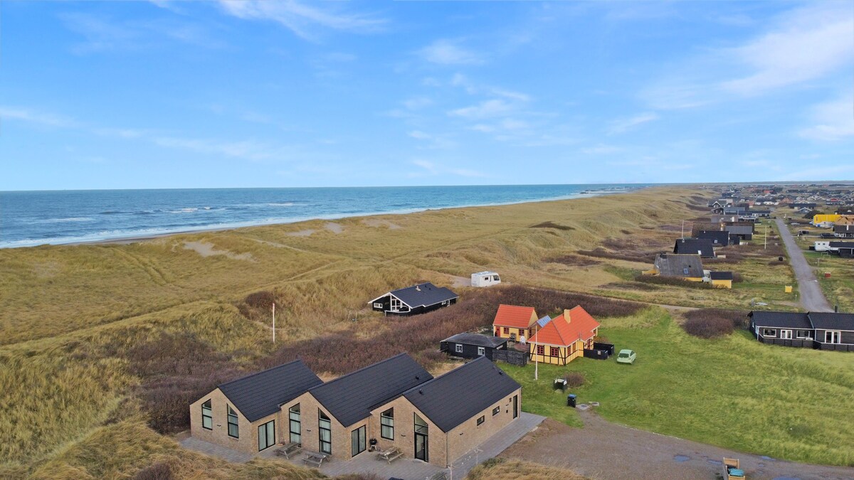 An aerial view captures multiple holiday homes set against a backdrop of sandy dunes and the North Sea. The coastline stretches in the distance, while various building styles and colors dot the landscape, showcasing the inviting setting for relaxation and outdoor activities.