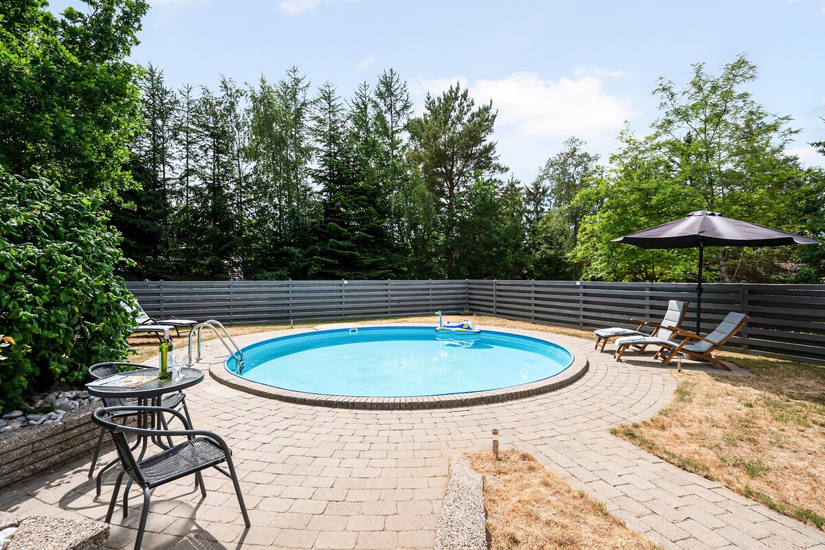 An inviting outdoor area features a round swimming pool surrounded by paved stones. Sun loungers are placed nearby, and an umbrella provides shade. Lush greenery borders the space, enhancing the sense of privacy. A few chairs are positioned on the patio for relaxation.