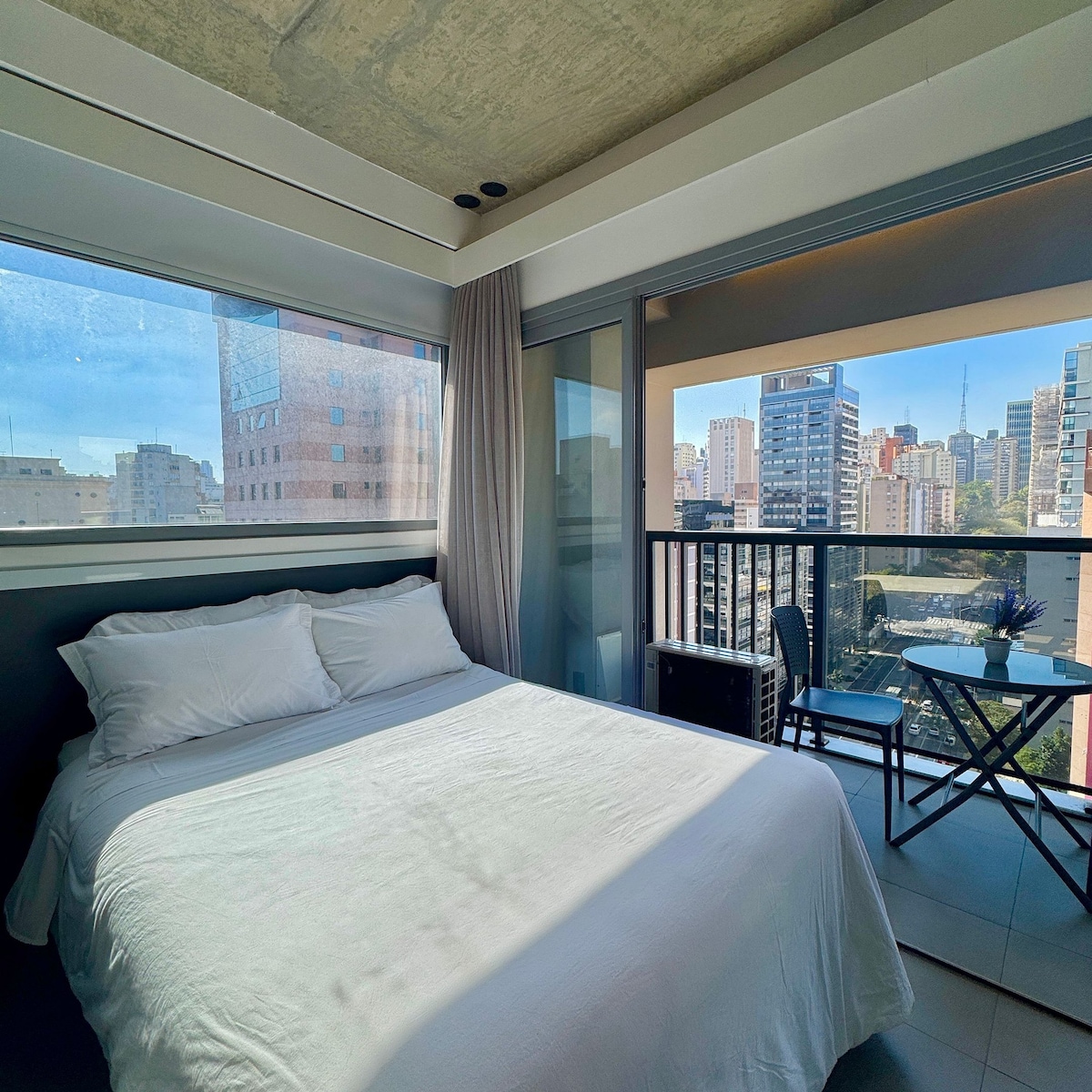 A bright bedroom is highlighted by large windows that provide city views. A spacious bed is dressed in crisp white linens, while a small balcony features a table and chair, inviting moments of relaxation.