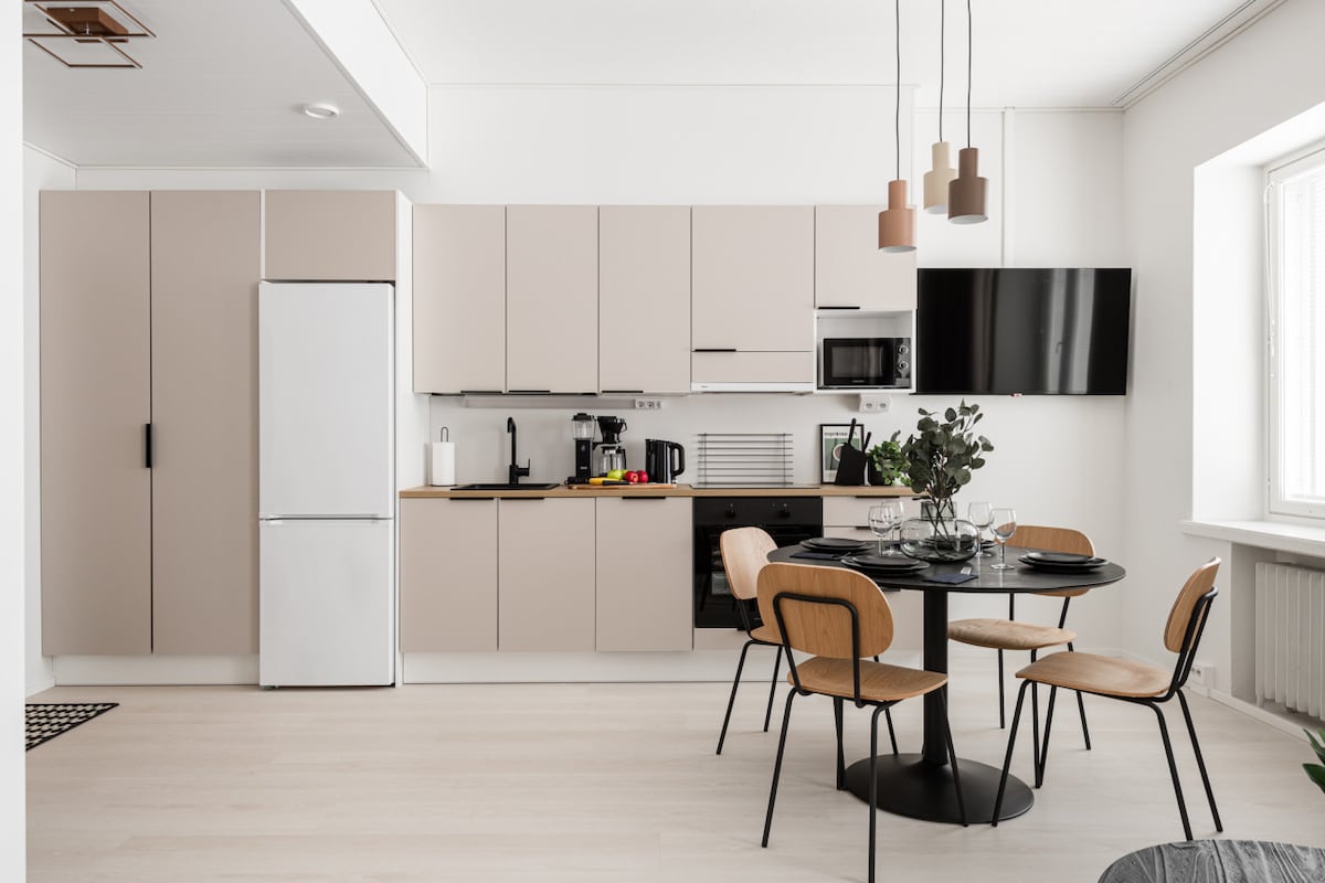 A modern kitchen area features sleek beige cabinetry and a spacious countertop. A dining table with four wooden chairs is set for meals, while a wall-mounted television is positioned above the kitchen appliances, which include a refrigerator, microwave, and coffee maker.
