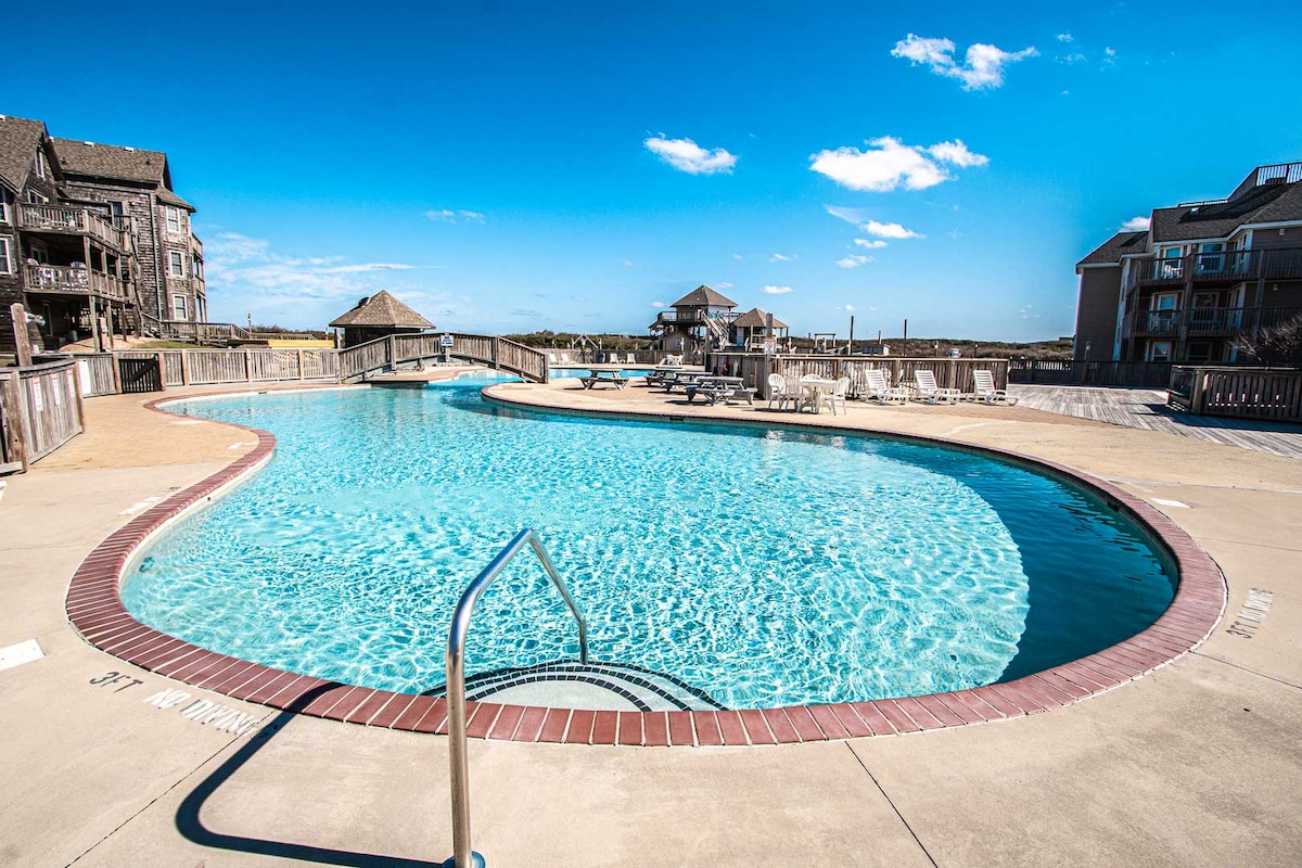 A spacious outdoor pool is seen, featuring a curved design and clear blue water. Surrounding the pool, a deck area includes sun loungers and tables. The backdrop consists of resort buildings and a clear sky, creating a relaxing outdoor atmosphere.