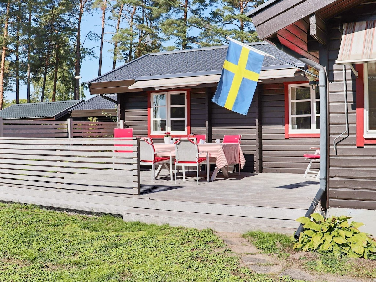A wooden terrace features a large dining table with several chairs, accompanied by a flag pole displaying the Swedish flag. The house exterior is dark wood, and other cottages are visible in the background, surrounded by grassy areas and trees.