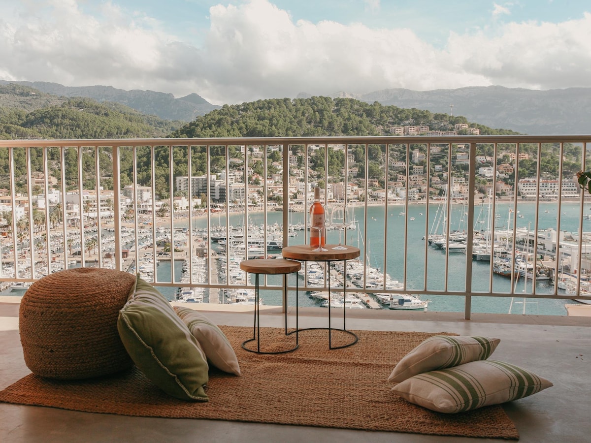 A terrace area is visually presented with two small tables and a cozy seating arrangement featuring a floor cushion. The view reveals the tranquil waters of Port de Sóller, lined with boats and surrounded by lush green mountains under a partly cloudy sky.