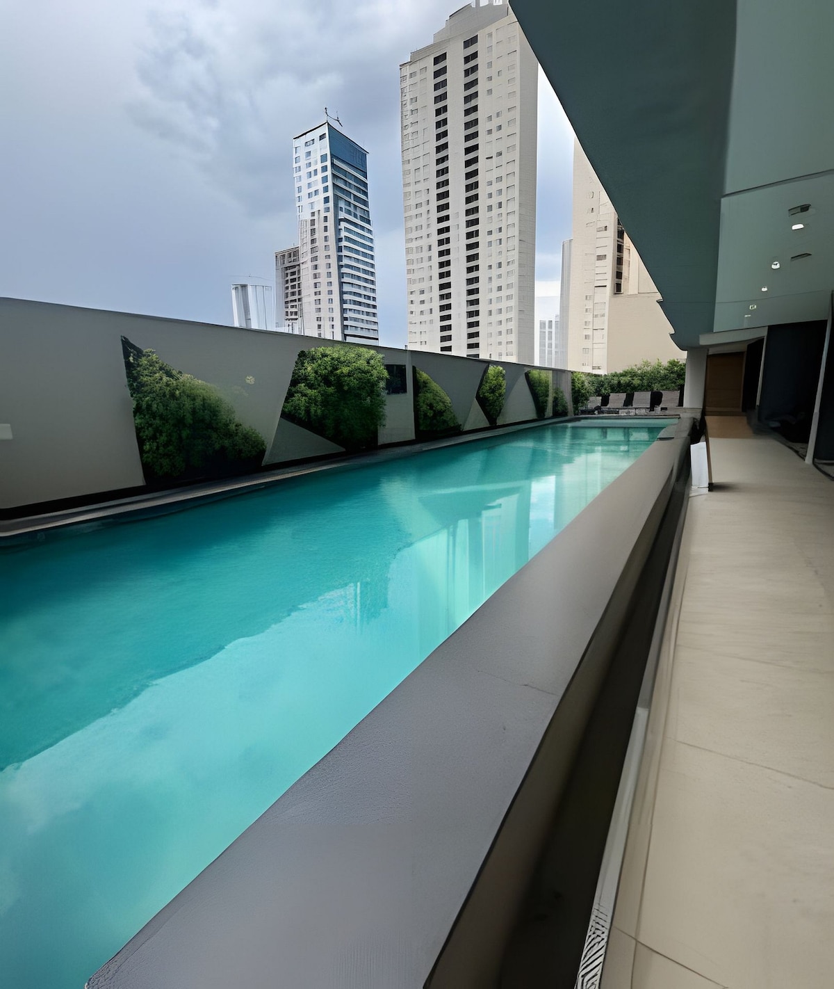 An outdoor pool is shown, reflecting the city skyline. Surrounding walls feature green landscaping, creating a contrast against the modern architecture. The pool area is well-lit, offering a sense of relaxation amidst the urban environment.