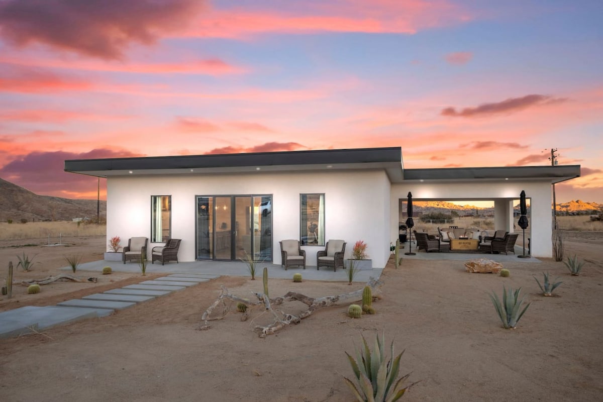 A modern single-story home is presented against a colorful sunset. The exterior features large glass doors that create a seamless view of the surrounding landscape. A spacious outdoor seating area is visible, furnished with comfortable chairs and a dining table, surrounded by desert plants.