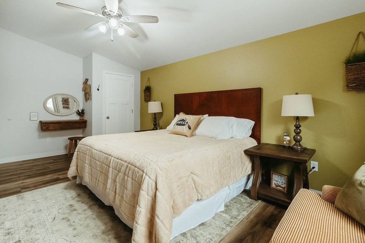 A spacious bedroom is equipped with a large bed featuring neutral bedding. Two bedside tables support lamps for ambient lighting. A decorative mirror hangs on the wall, and a cozy seating area with an accent chair is present. A warm yellow wall adds a touch of color.