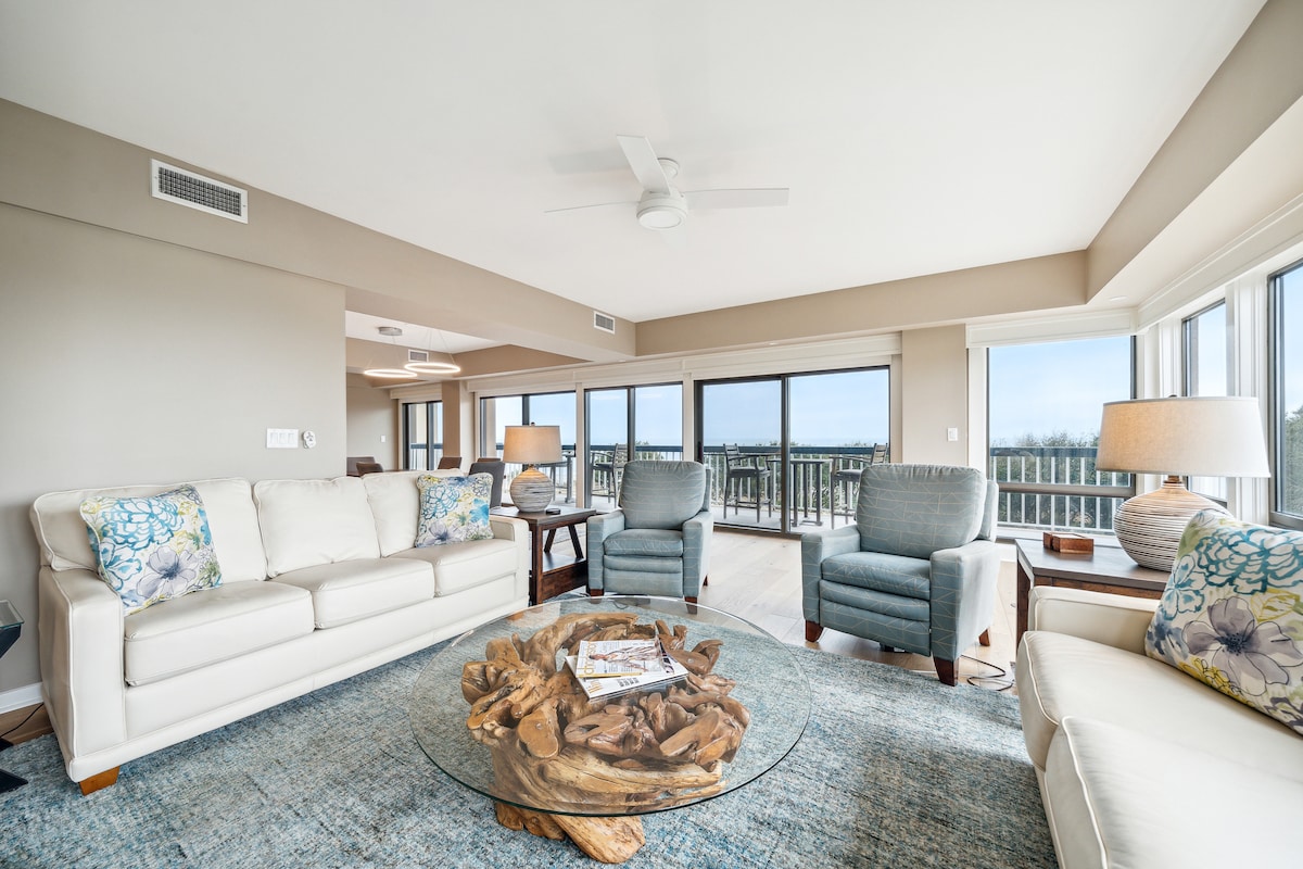 A bright living area features a large, circular glass coffee table set atop a soft area rug. Light-colored sofas and blue armchairs provide comfortable seating, with large windows offering scenic views. A ceiling fan enhances air circulation in the spacious room.