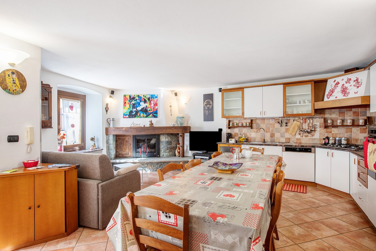 An open-plan living area is featured, showcasing a long dining table surrounded by wooden chairs. A comfortable sofa is arranged adjacent to a fireplace. The kitchen is fully equipped with cabinetry and appliances, creating a warm and functional space for guests.