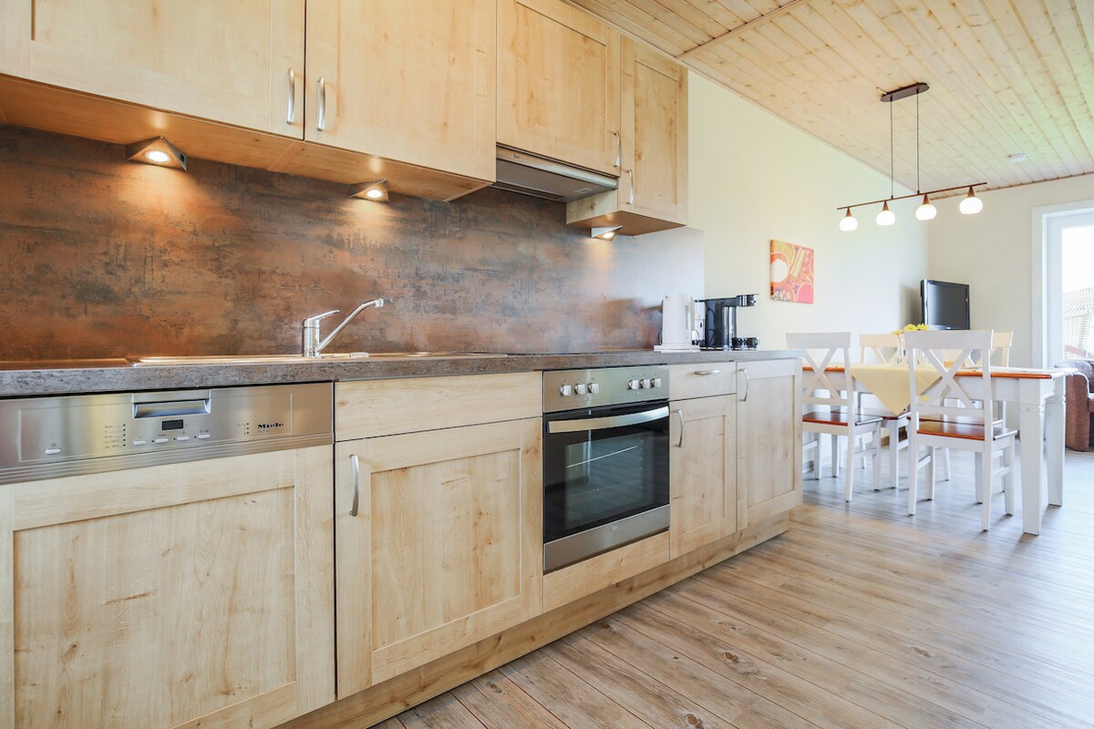 A modern kitchen features wooden cabinetry and stainless steel appliances, including an oven, stovetop, and dishwasher. Soft lighting highlights a spacious countertop. An adjacent dining area with a light wooden table and white chairs is in view, complementing the open layout of the living space.