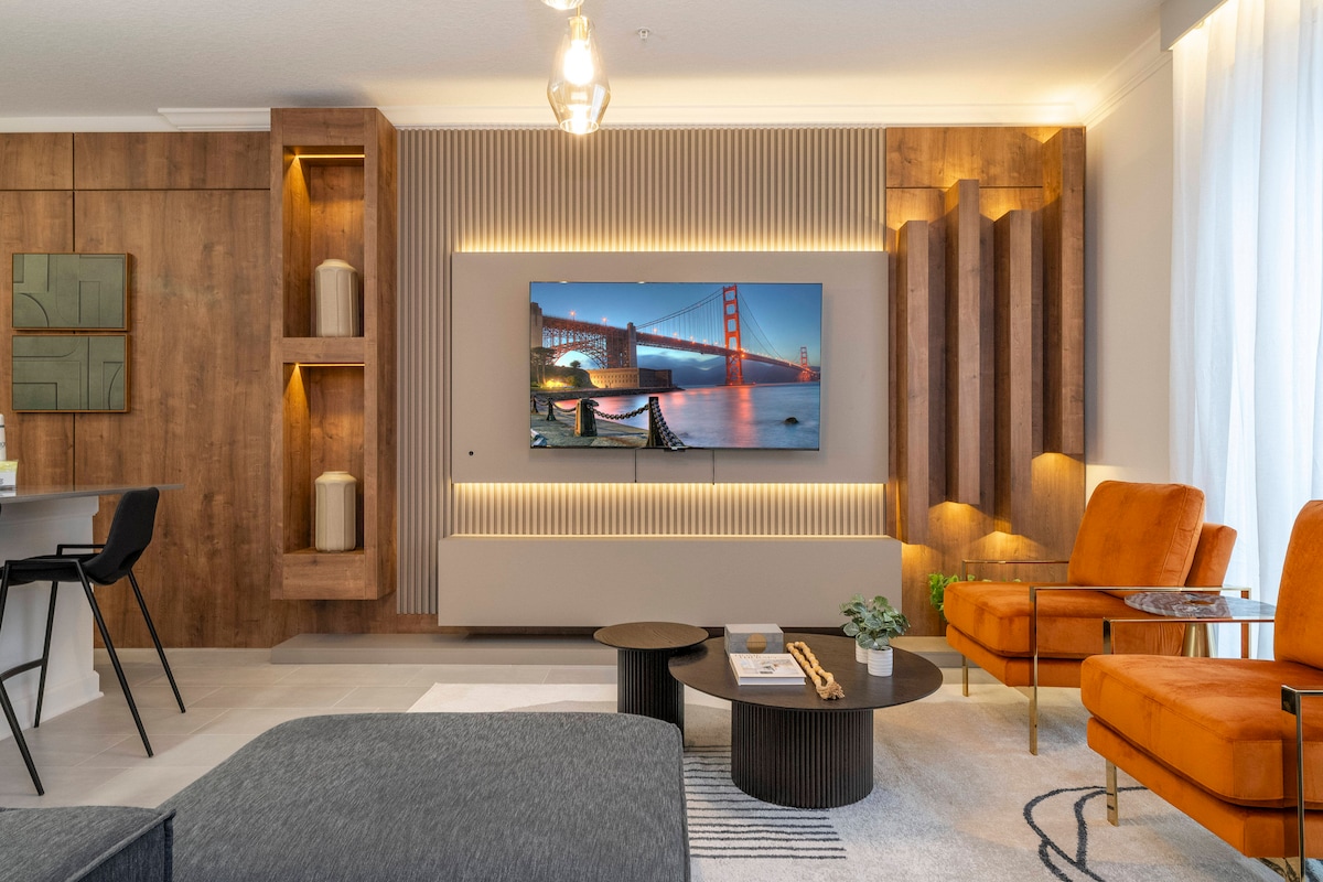 A modern living area is showcased, featuring a sleek sofa and two orange armchairs. A large television is mounted on a textured wooden wall, displaying a cityscape. Subtle lighting accents the decor, while a round coffee table is positioned at the center, accompanied by a small plant.