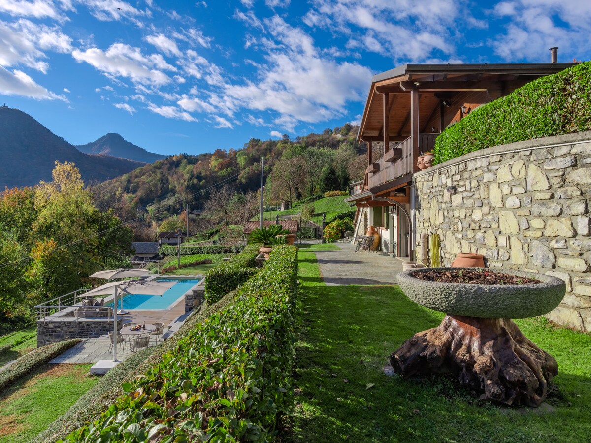 The chalet is set against a backdrop of rolling hills, surrounded by lush greenery. A swimming pool reflects the sky, and a stone fountain complements the landscaped garden. Sunlight illuminates the outdoor area, enhancing the tranquil ambiance of the property.
