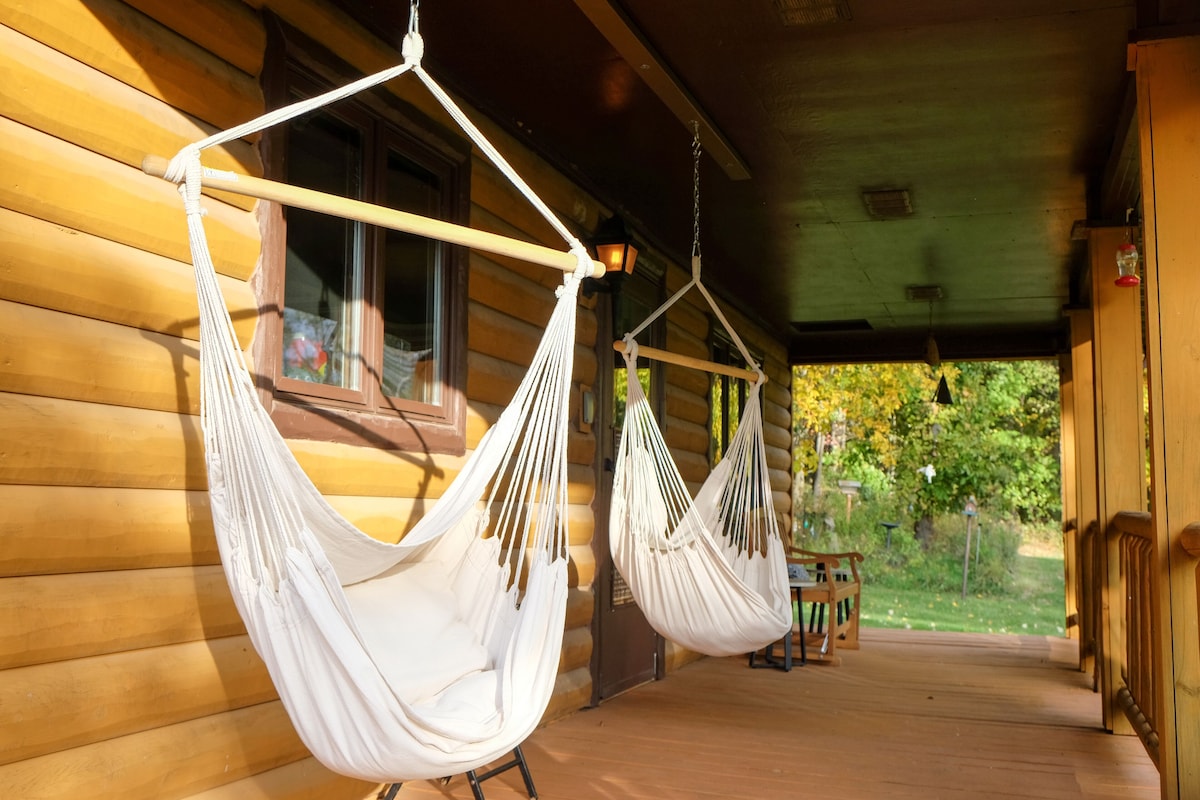 Two hanging hammock chairs are positioned on a covered porch, offering a relaxed seating area. The warm wooden tones of the cabin exterior complement the natural surroundings. Soft lighting from the interior enhances the inviting atmosphere, while greenery is visible in the background.