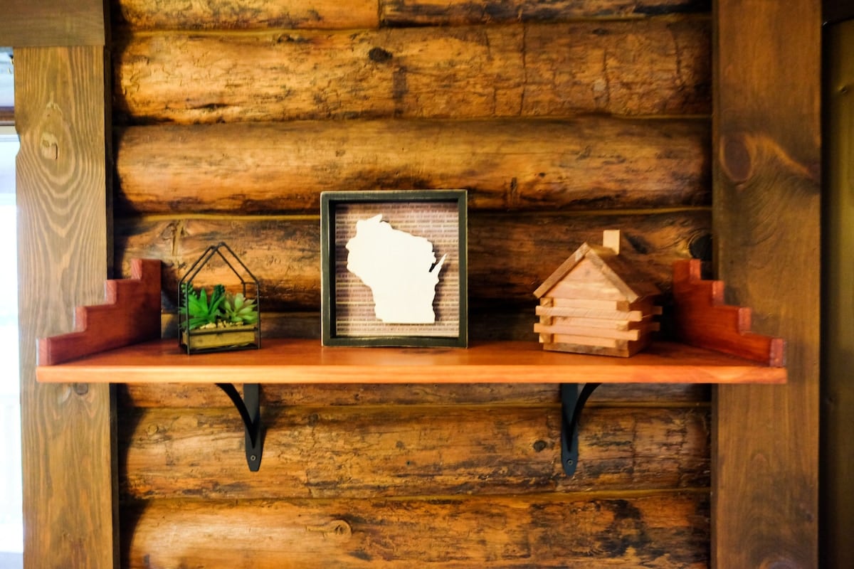 A wooden shelf is mounted against a backdrop of rustic log walls. Decorative items include a framed outline of Wisconsin, a miniature log cabin, and a small glass terrarium with greenery, adding a touch of nature to the space.