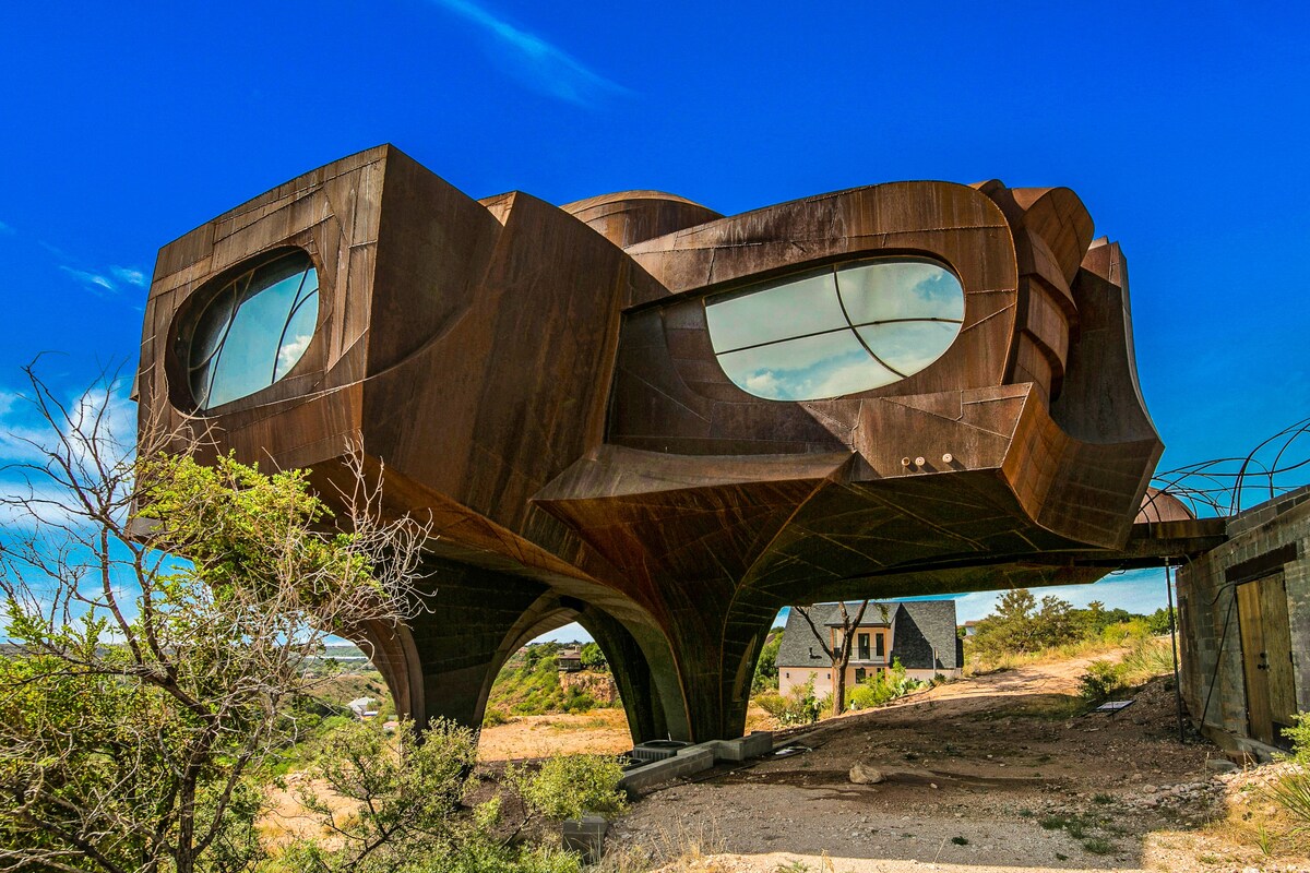 An architecturally unique house is suspended on slender supports, featuring large, rounded windows that reflect the sky. Its rust-colored exterior showcases intricate, sculptural designs against a backdrop of natural landscape, highlighting the distinctive structure's artistic appeal.