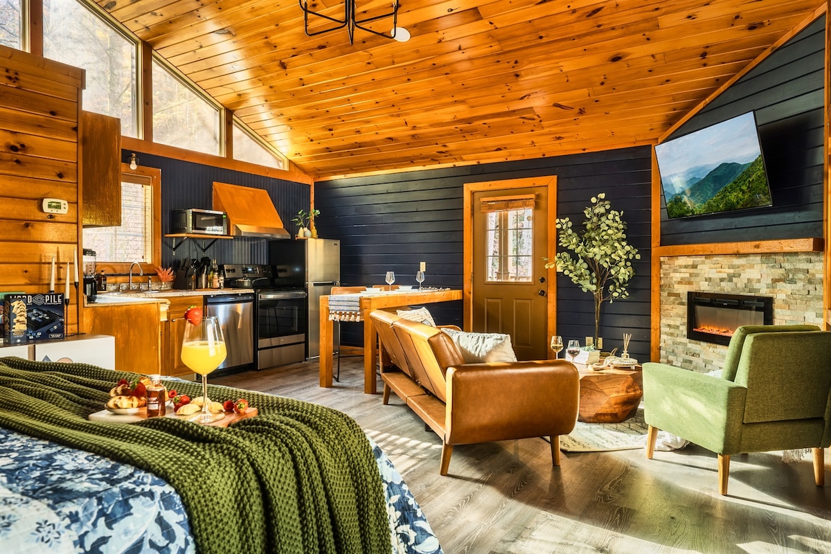 An open living area is highlighted by a vaulted wooden ceiling and large windows allowing natural light. A kitchen area with modern appliances is visible alongside a dining space. Comfortable seating options include a green armchair and a brown sofa, accented by decor and an electric fireplace.