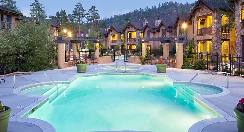 A serene outdoor heated pool is surrounded by lush greenery and elegant stone buildings, illuminated by soft lights at dusk. The pool features steps leading into the water, with comfortable seating areas nearby for relaxation.
