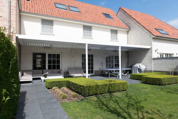A spacious outdoor area features a covered patio with comfortable seating and a dining space. Well-maintained hedges border the garden, which includes a manicured lawn. Sunlight streams down, highlighting the charming brick façade of the house and the inviting atmosphere of the space.
