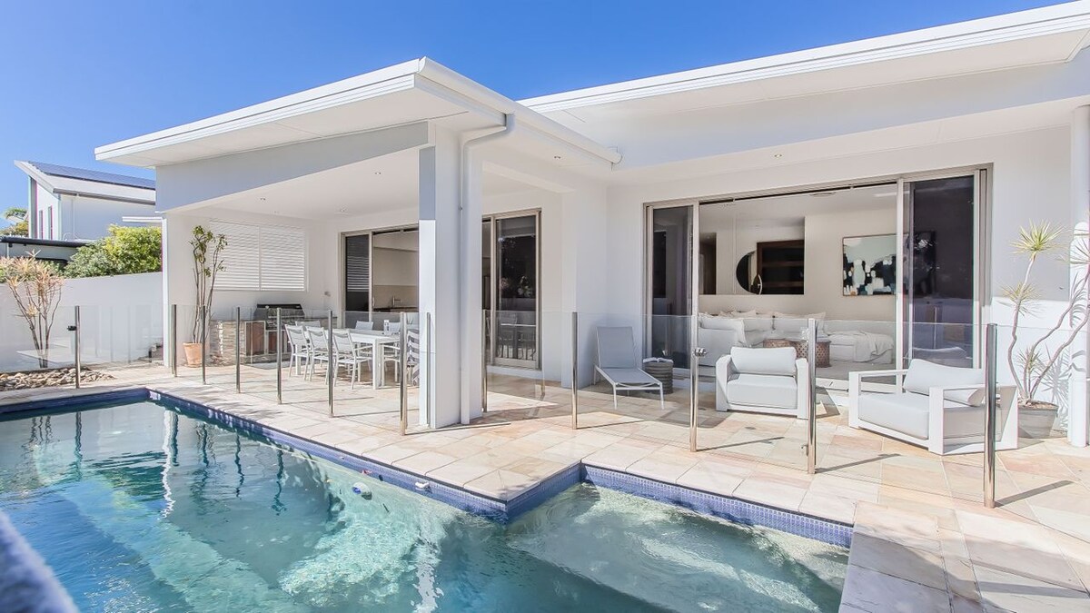 A serene outdoor area features a private pool surrounded by sunlit stone tiles. Stylish lounge chairs are positioned near the pool, with an outdoor dining table visible beyond. Large sliding glass doors provide a seamless connection to the indoor living space.