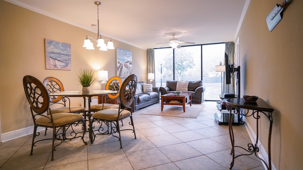 An open living area features a round dining table with four chairs, surrounded by light-colored walls. A comfortable seating arrangement includes a dark couch and coffee table, while large windows provide ample natural light and a view of the outdoors.