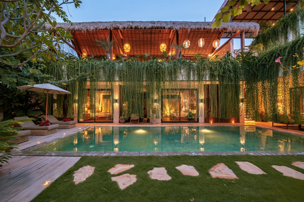 The outdoor area showcases a tranquil swimming pool surrounded by lush greenery and vibrant foliage. Warm light from the overhead fixtures adds to the inviting ambiance, while a stylish patio leads to an open living space, completing the tropical living experience.