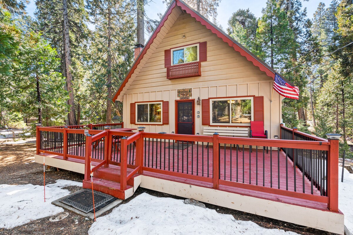 A rustic cabin features a warm wooden exterior with a slanted roof and a welcoming porch. A red railing surrounds the deck, providing an inviting entryway. American flags adorn the front, and large windows allow natural light to fill the space. Snow covers the ground in the surrounding area.