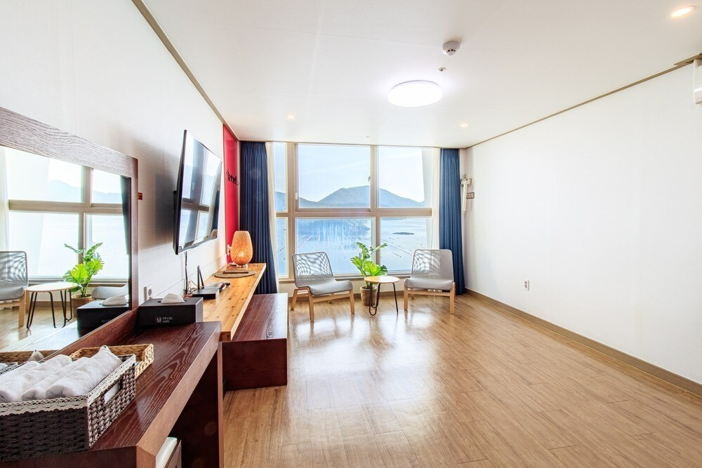A bright living area features large windows offering a panoramic ocean view. Two seating areas are arranged with light chairs, accompanied by potted plants. A flat-screen TV is mounted on the wall, while a wooden console holds decorative items and a small basket.