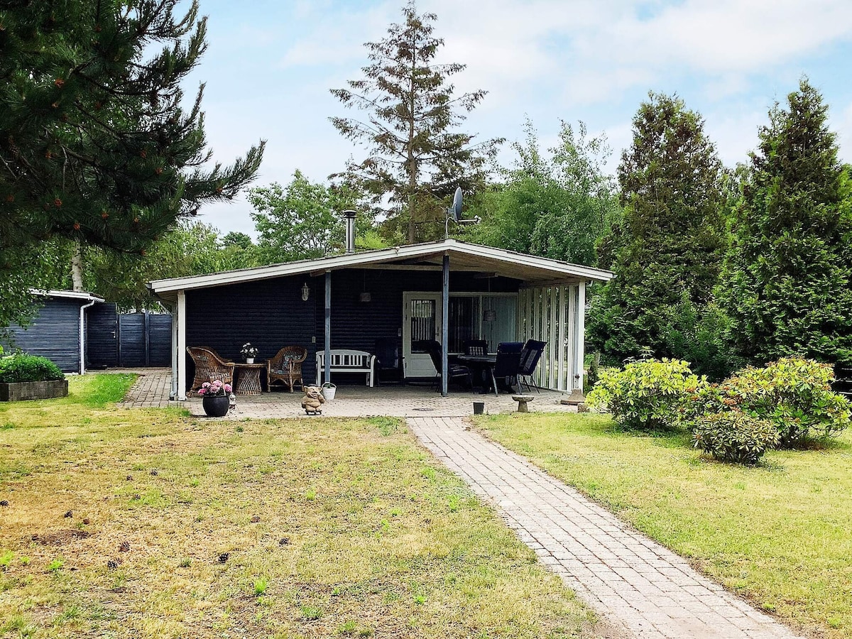 The exterior of a holiday home is displayed, featuring a covered terrace with seating arrangements. A paved pathway leads through a garden area adorned with greenery and trees. The relaxed setting invites enjoyment of the surrounding nature.