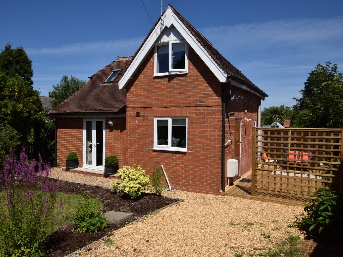 A charming brick cottage is shown, designed with a steeply pitched roof and multiple windows. Flowering plants and a gravel path enhance the garden space, which features a wooden fence leading to a small patio area with outdoor seating.