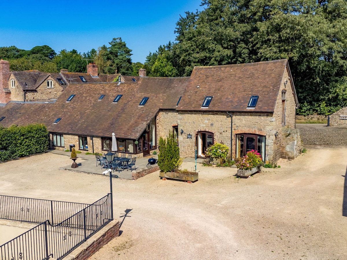A charming stone building is set in a rural landscape. The property features a welcoming patio area with outdoor furniture and flowering plants, surrounded by gravel paths. Sunlight filters down, highlighting the rustic architecture and natural surroundings.
