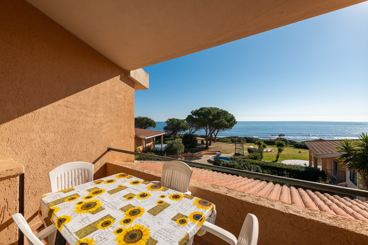 A balcony view is presented, featuring a table adorned with a sunflower-patterned tablecloth. The serene blue sea and clear sky are visible in the background, complemented by lush greenery in the foreground. The scene invites relaxation and appreciation of the coastal surroundings.