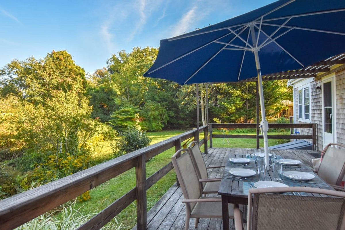 A spacious deck houses a large dining table set with plates and glasses, complemented by comfortable seating. A large umbrella provides shade, while the lush green yard and trees enhance the outdoor setting, creating an inviting space for relaxation and dining.
