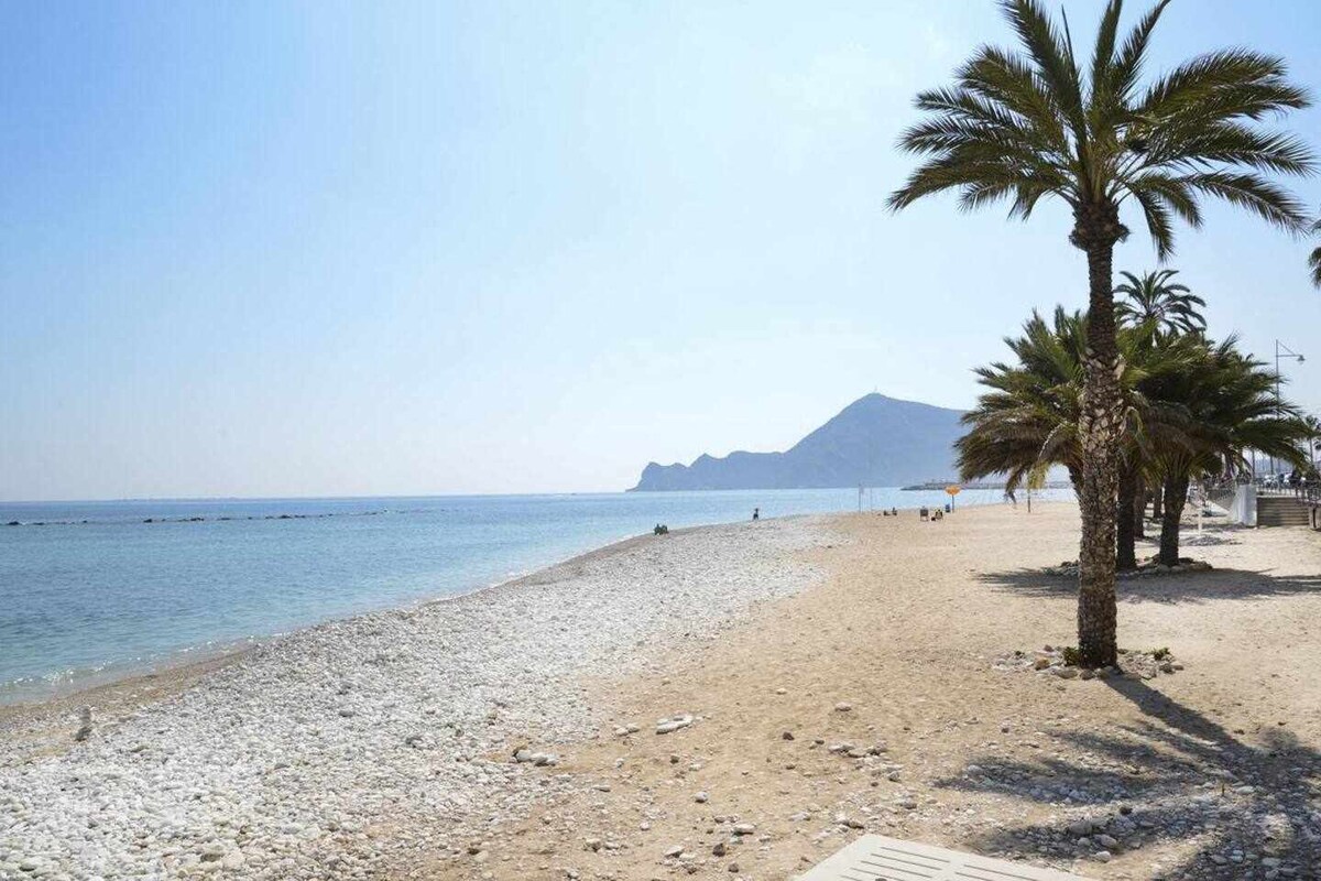 A serene beach scene features a gentle shoreline lined with palm trees. Soft sands blend with smooth stones along the water's edge, leading to clear blue waters. In the background, a distant mountain silhouette is visible against a clear sky.