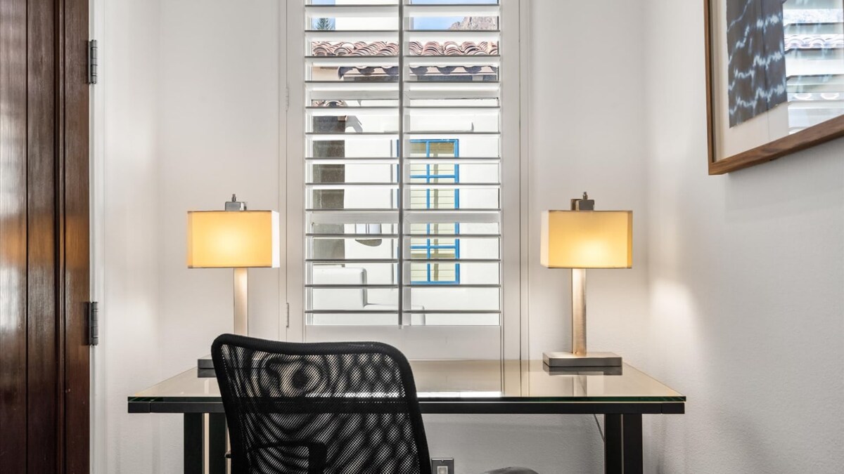A sleek workspace is presented with a glass-top desk accompanied by two modern lamps. A comfortable chair is positioned in front of the desk, while natural light filters through a shuttered window, offering a view of the exterior.
