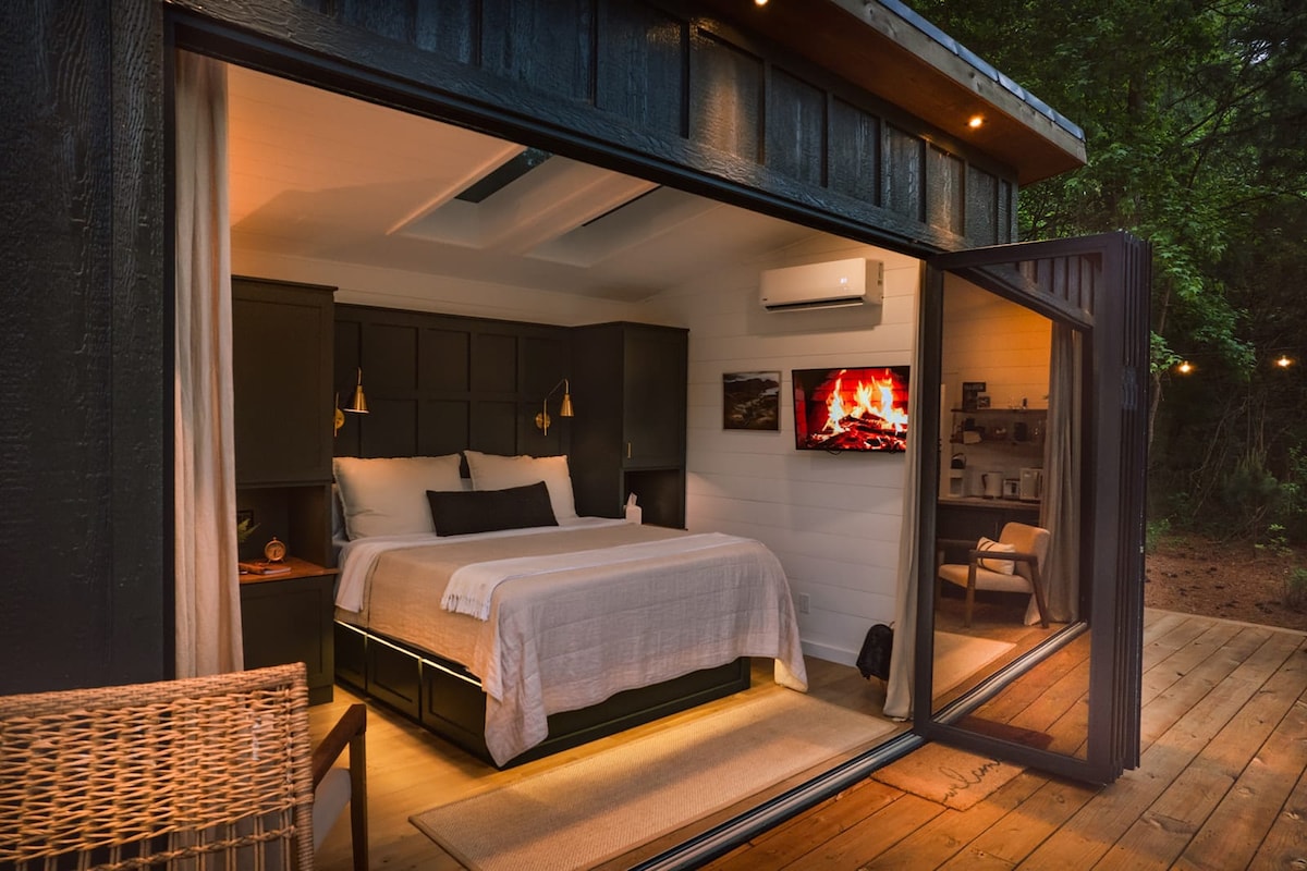 A modern cottage room is visible, featuring a spacious bed with a neutral-colored blanket. Two bedside lamps provide soft lighting. A wall-mounted television displays a fireplace image. Large glass doors open to a wooden deck, surrounded by trees, with a wicker chair positioned outside.