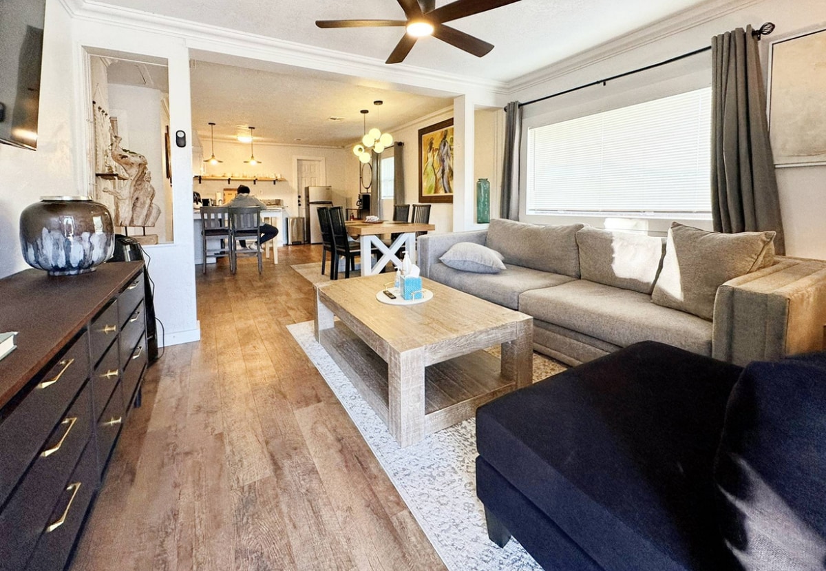 An inviting living room features a large sectional sofa and a coffee table at its center, complemented by a stylish area rug. Natural light streams through a window, illuminating the space, while a ceiling fan and wall-mounted television enhance the modern ambiance.