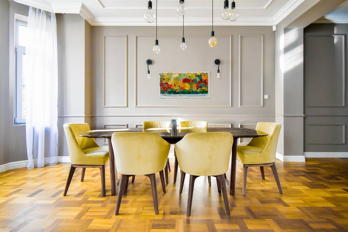 A dining area features a large circular table surrounded by six yellow upholstered chairs. Warm wood flooring is highlighted by natural light filtering through a window. The soft gray walls are complemented by a colorful artwork piece above the table.