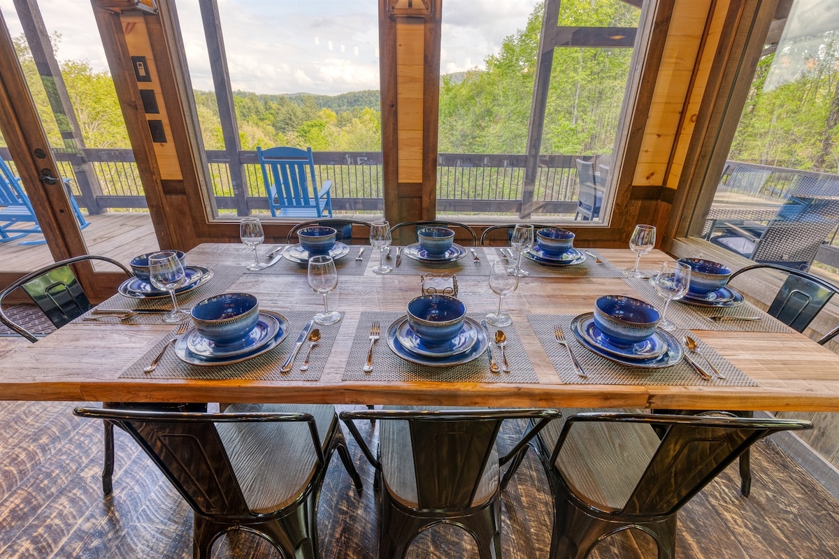A spacious dining table is set for multiple guests, featuring blue dishware and glassware. Tall windows reveal a wooded landscape outside, allowing natural light to brighten the interior. Chic black chairs surround the table, creating a modern dining environment.