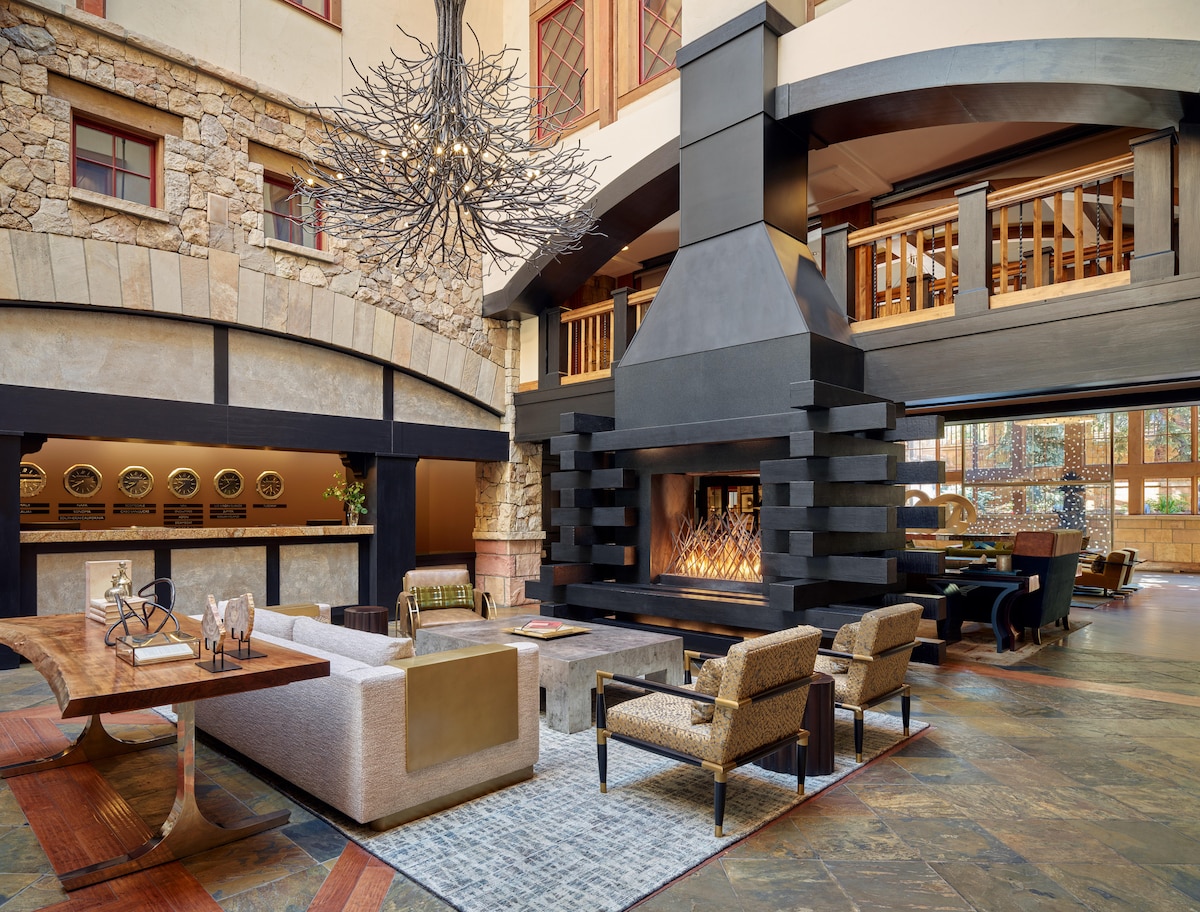 A spacious lobby area features a large stone fireplace at its center, surrounded by comfortable seating. A unique chandelier hangs overhead, and natural light streams through expansive windows. Rich wood accents complement the diverse textures of stone and furniture throughout the space.