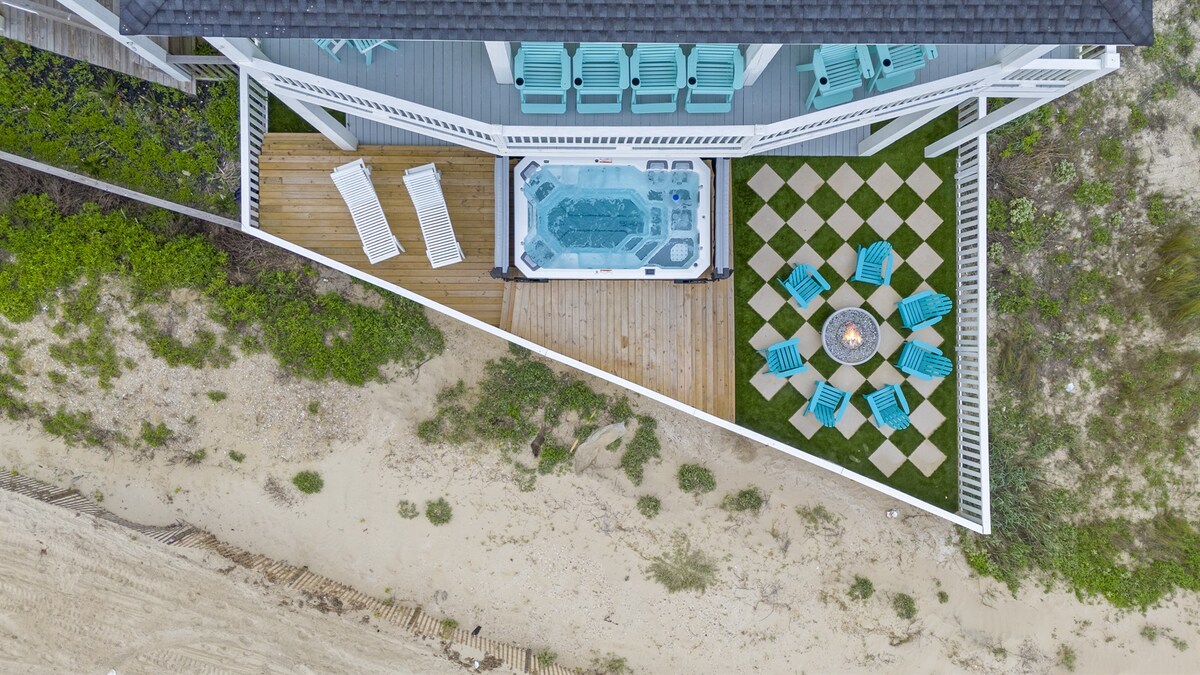 An aerial view showcases the outdoor area featuring a SwimSpa and a fire pit surrounded by turquoise seating. A sun deck with lounge chairs complements the vibrant outdoor space, while the sandy beach is visible just beyond the landscaped turf backyard.