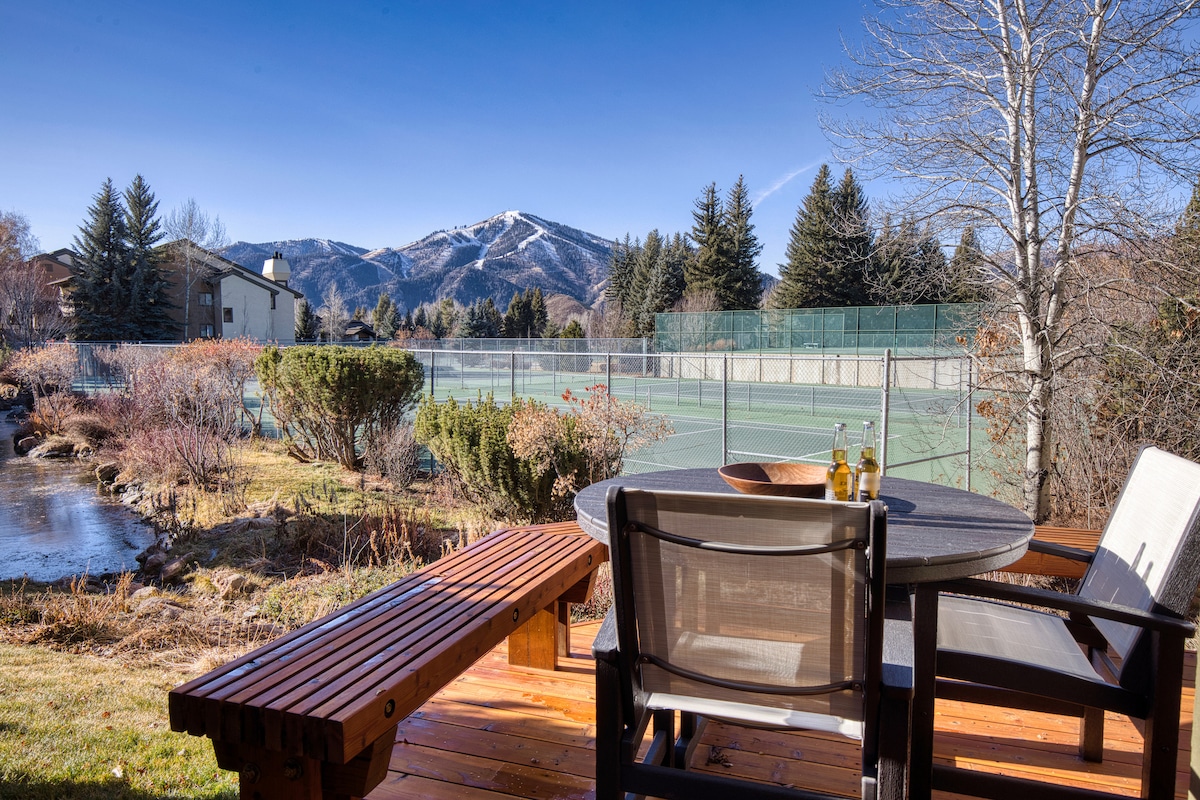 A private deck features a circular table surrounded by comfortable chairs. The backdrop displays well-maintained tennis courts and lush landscaping, set against the majestic mountain views under a clear blue sky.