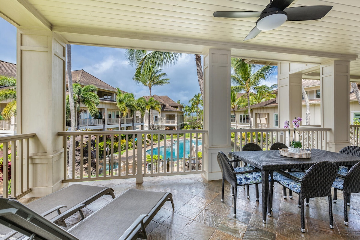 An outdoor lanai is furnished with a dining table and chairs, surrounded by tropical landscaping. Two loungers are positioned nearby, offering views of the resort-style pool and palm trees swaying gently in the breeze. The space is designed for relaxation and enjoyment of the outdoors.