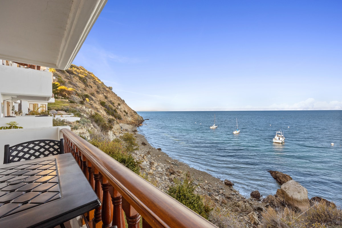 A balcony view reveals gentle ocean waves lapping against the rocky shoreline. Several boats are anchored in the tranquil water, framed by the surrounding hills under a clear blue sky.