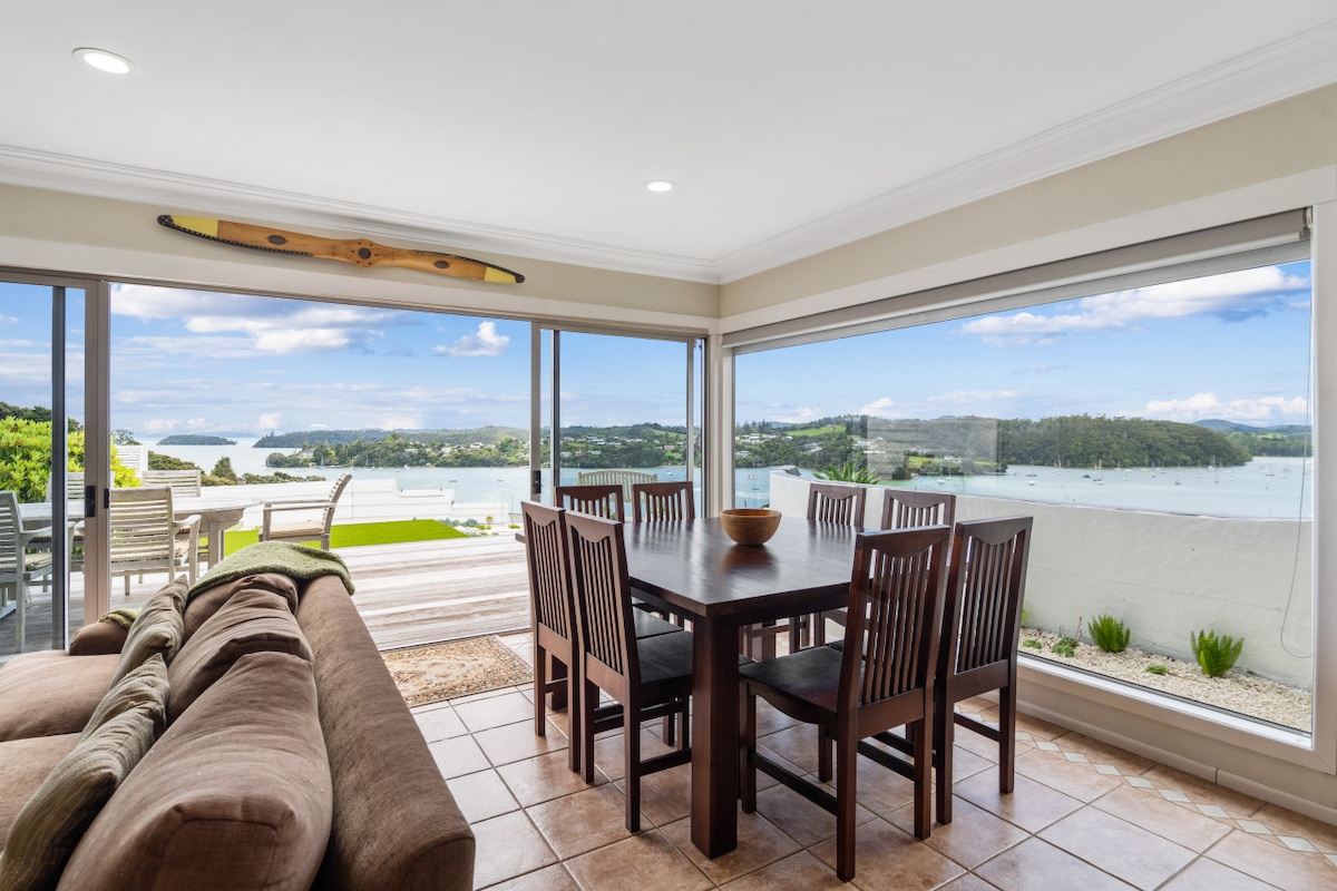 A dining area features a large wooden table surrounded by eight chairs, with natural light flooding in from expansive windows. A comfortable seating arrangement is positioned nearby, offering views of the Opua Marina and harbor, enhancing the relaxed atmosphere of the space.