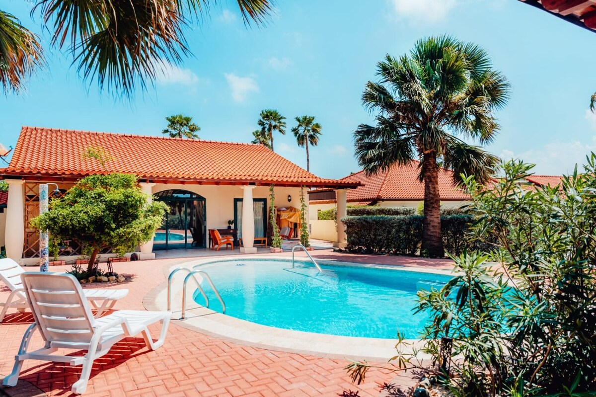 A large private pool is surrounded by palm trees and lush greenery. The area features lounge chairs arranged along the poolside, and a tiled patio enhances the outdoor space. The inviting pool reflects the clear blue sky above, creating a tranquil atmosphere.