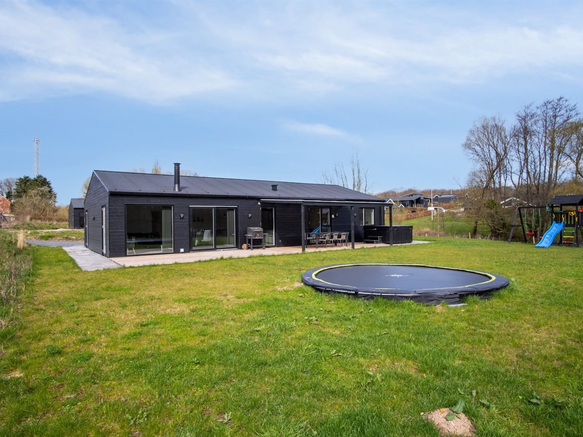 The exterior features a modern black single-family house with expansive glass windows and a flat roof. A spacious lawn surrounds the property, with a round trampoline situated prominently in the foreground. A patio area is visible, offering space for outdoor seating.